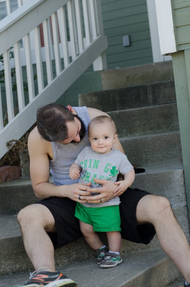 Finn, standing in front of daddy and leaning against him while he smiles and looks at the camera.